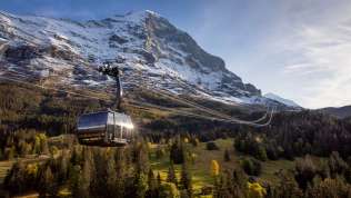 Die Eiger Express-Seilbahn im Frühling, mit grüner Landschaft im Vordergrund, schneebedeckten Bergen im Hintergrund und blauem Himmel.